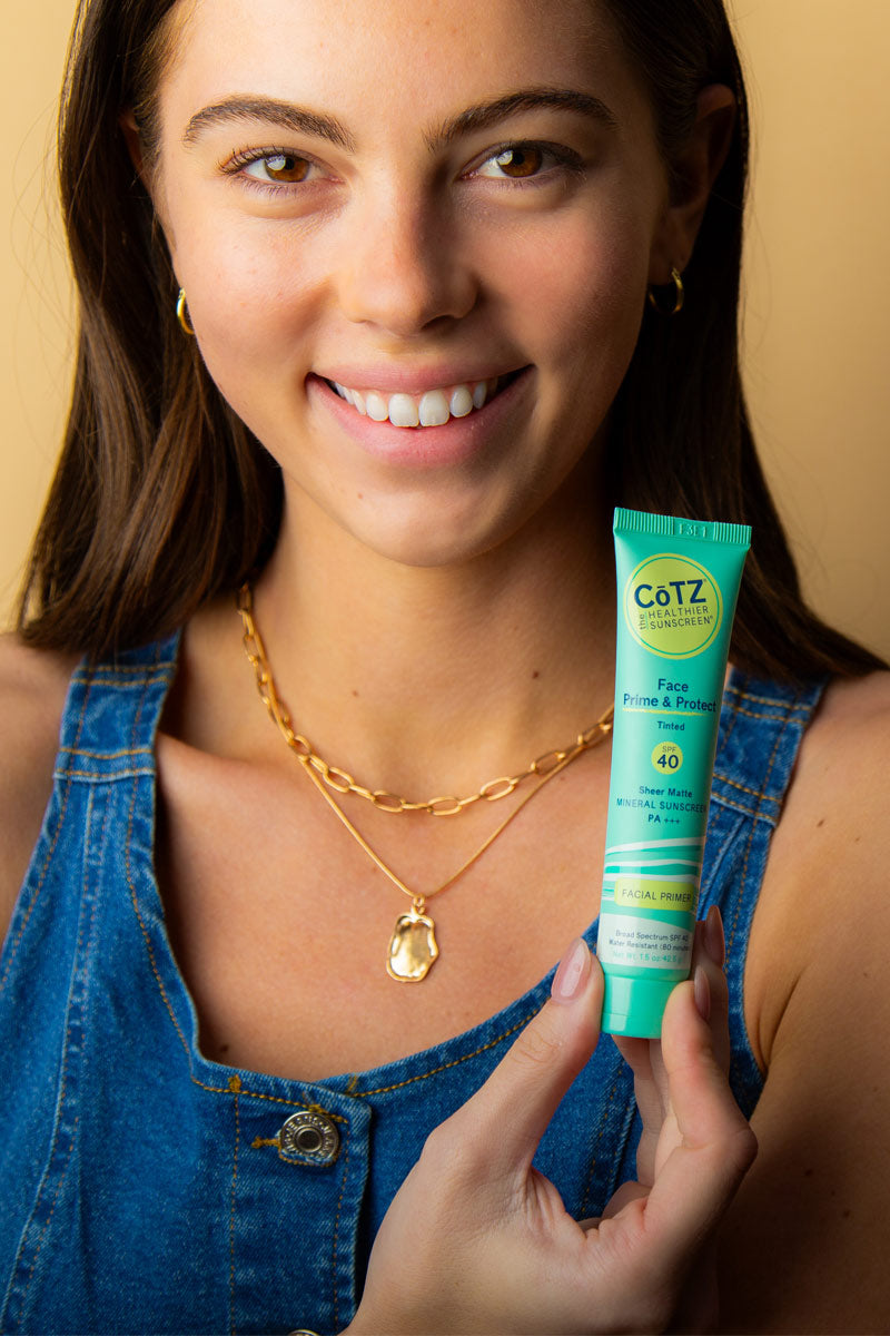 Image of model smiling holding Cotz Face Prime & Protect Tinted SPF 40 at shoulder height