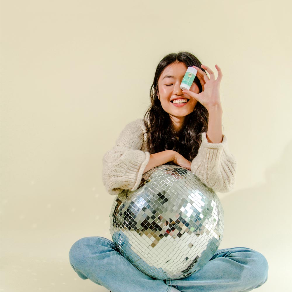 Model holding Cotz Mineral Sun Stick Tinted SPF 45 close to her eye while hugging a disco ball in her lap that is reflecting off light