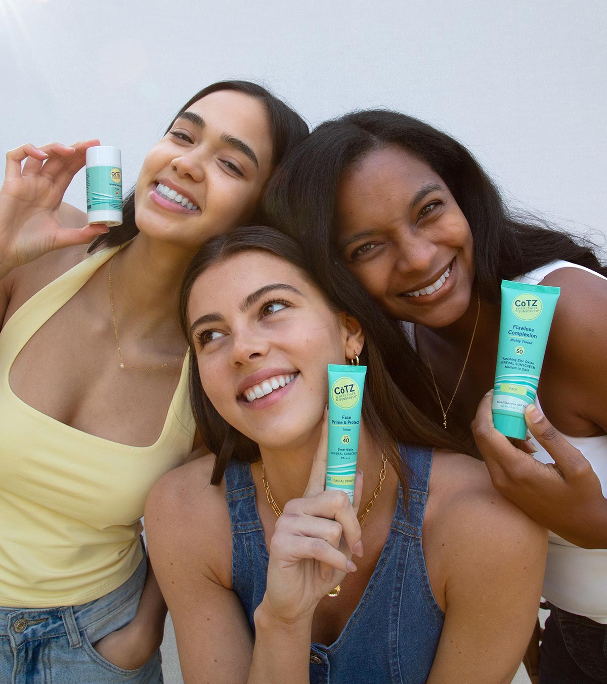 Three models smiling holding three different products: Cotz Mineral Sun Stick, Cotz Face Prime & Protect Tinted, and Cotz Flawless Complexion Richly TInted.