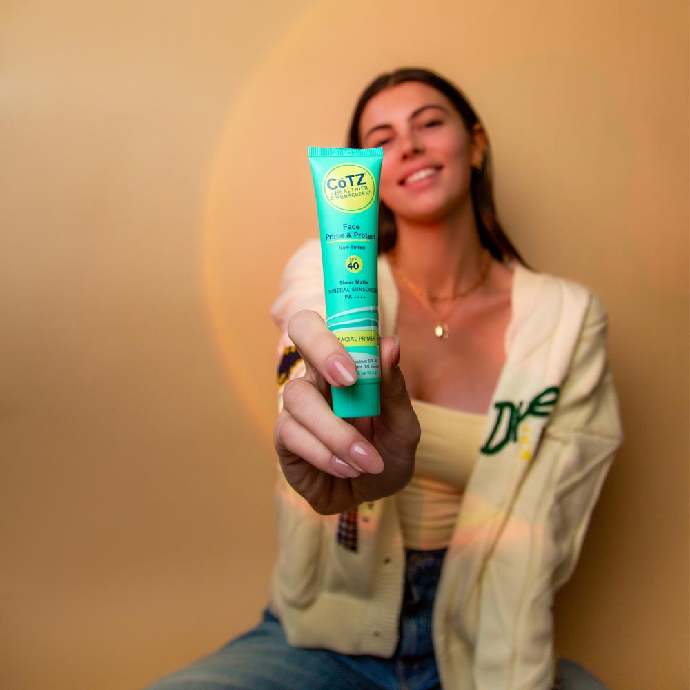 Model holding Cotz Face Prime & Protect Non-Tinted SPF 40 in front of her while smiling