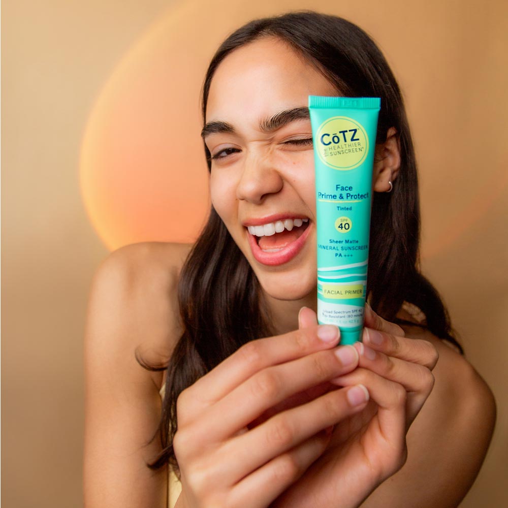 Image of model holding tube of Cotz Face Prime & Protect Tinted SPF 40 while winking