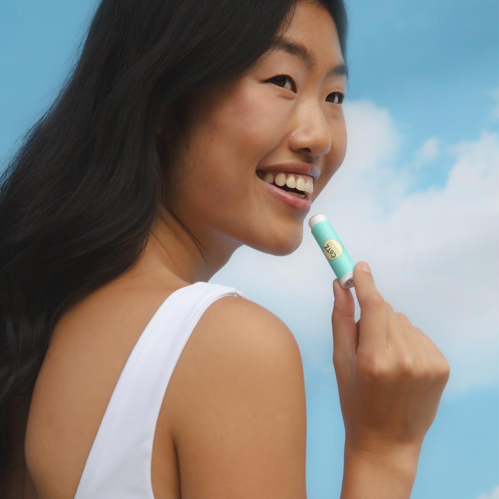 Image of a model holding and applying Cotz Lip Balm Tinted SPF 45 with the sky in the background