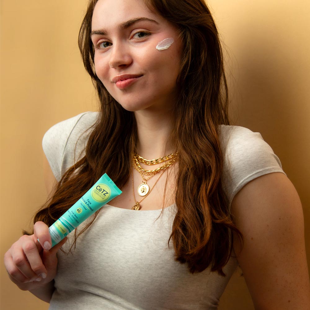 Model holding Cotz Face Prime & Protect Non-Tinted SPF 40 tube with a swatch on her cheek