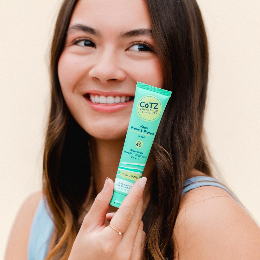 Image of model holding Cotz Face Prime & Protect Tinted SPF 40 tube near her face while smiling